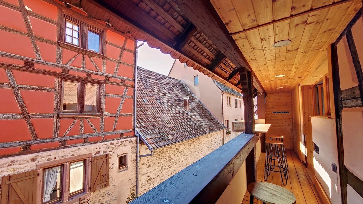 An outdoor terrace area shows wooden flooring with a bar-style seating arrangement. The view includes charming half-timbered buildings and rooftops, framed by an overhanging wooden ceiling. Natural light fills the space through the open ceiling, creating a warm, inviting environment.