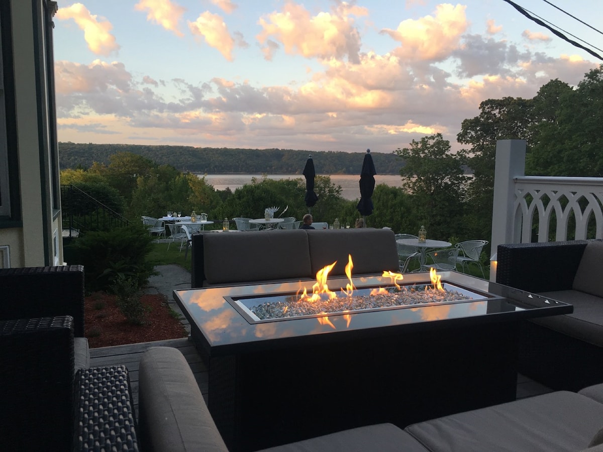 An outdoor seating area features a gas fire pit surrounded by modern lounge furniture. The scenic view includes rolling hills and a body of water in the distance, with clouds reflecting soft hues in the evening sky. Tables and chairs are positioned on the lawn for additional seating.