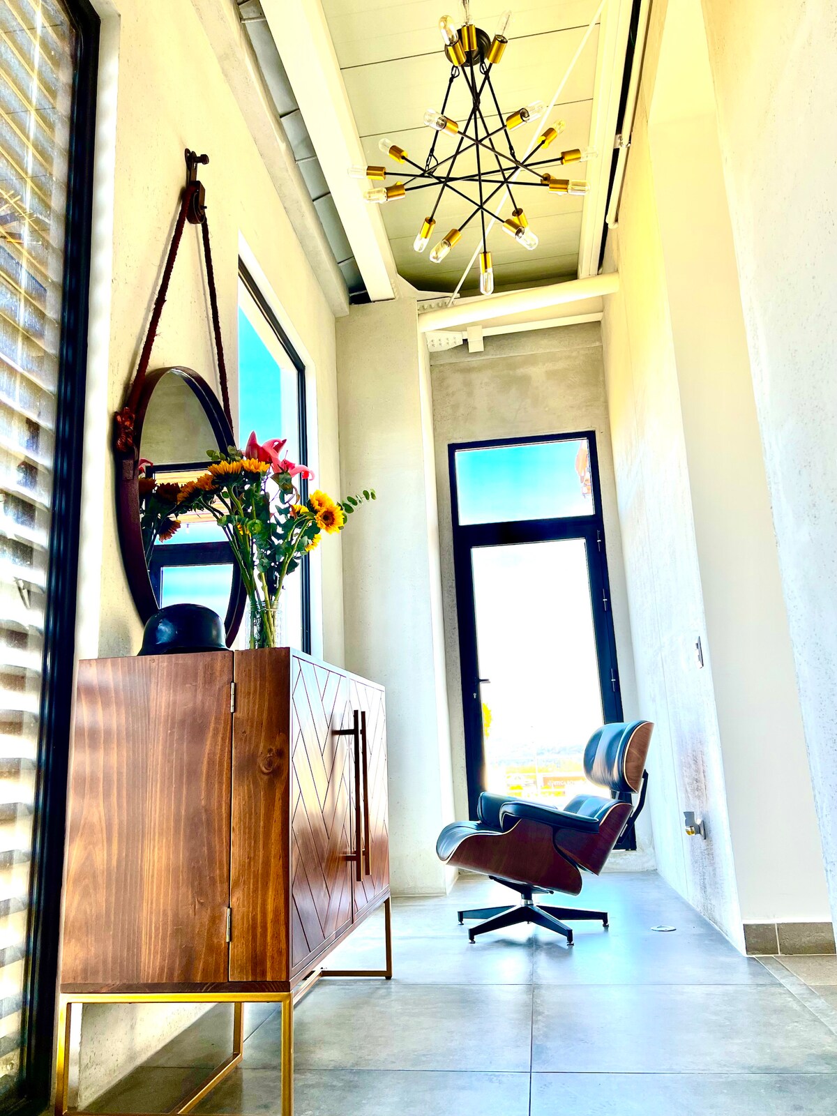 A modern hallway features high ceilings and large windows allowing natural light to flood the space. A stylish wooden cabinet contrasts with the neutral walls, accompanied by a sleek chair and a round mirror. Fresh flowers add a touch of color.