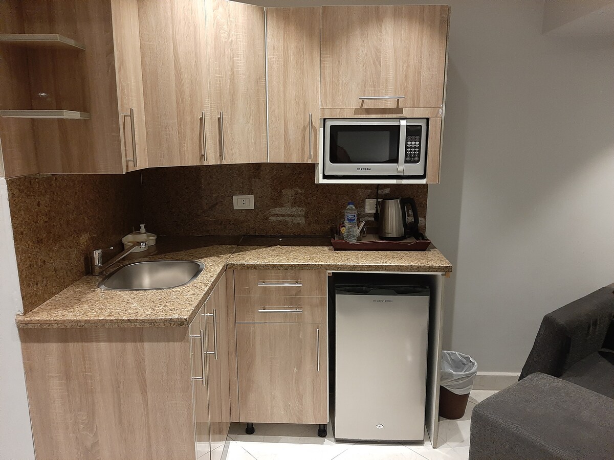 A kitchenette features wooden cabinetry and a modern countertop. A compact sink and mini refrigerator are visible, alongside a microwave and electric kettle. The area is well-organized, and neutral tones create a functional space for meal preparation.