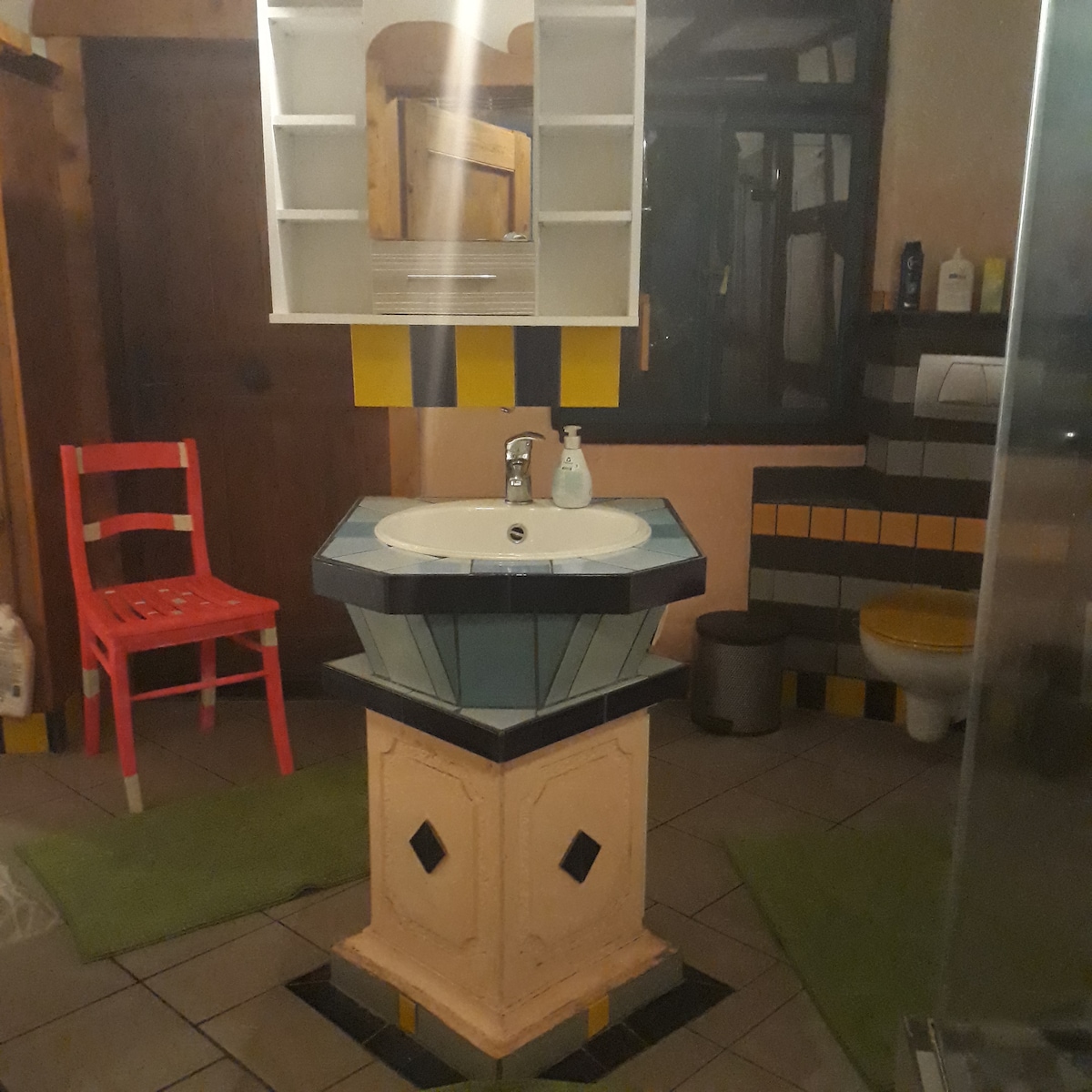 A unique bathroom features a pedestal sink with a geometric design, complemented by a large, framed mirror above it. Soft green mats are positioned on tiled flooring. A bright red chair adds a pop of color, while a shower area is visible in the corner.
