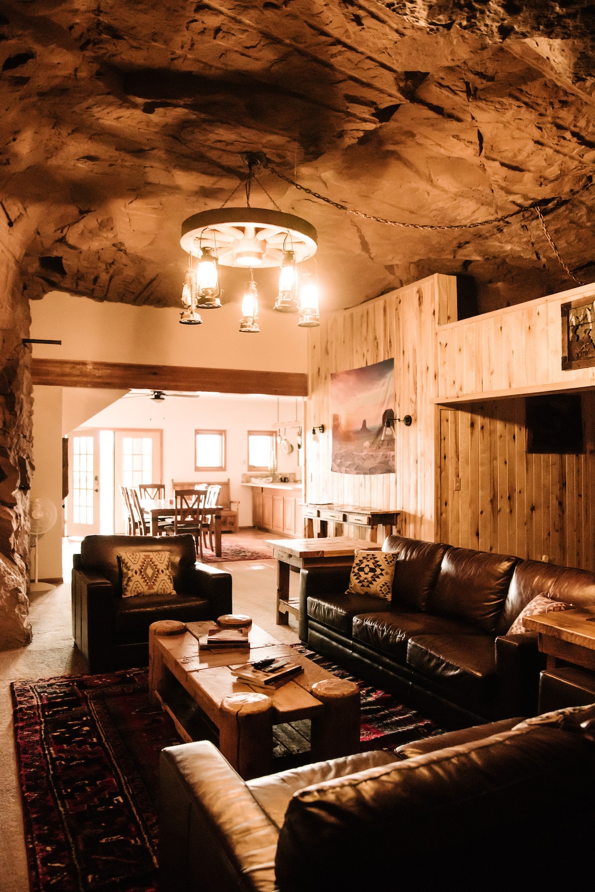The main living area features a rustic design, with exposed red rock walls and warm wood paneling. Two leather sofas provide comfortable seating, complemented by decorative pillows. A wooden coffee table sits at the center, surrounded by a vibrant area rug. Soft lighting enhances the inviting space.