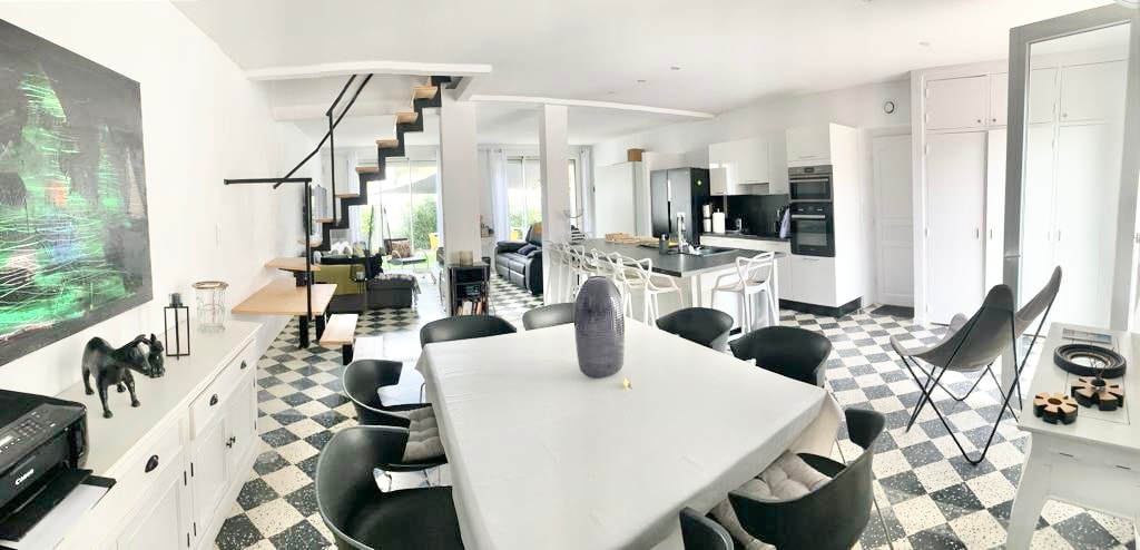 An open-concept living area features a large dining table surrounded by black chairs. A modern kitchen is visible in the background, with sleek appliances. A staircase is positioned to one side, leading to a second level. Natural light fills the space through expansive windows.