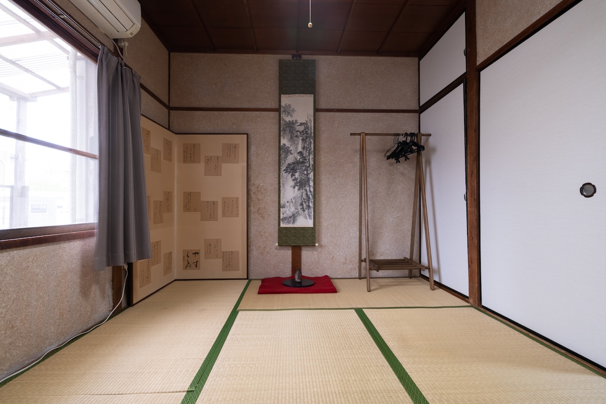 A traditional Japanese room features tatami flooring with a red accent mat. A decorative scroll hangs on the wall, complemented by a folding screen. A wooden coat rack is positioned next to the window, which is covered with light curtains, allowing natural light to fill the space.