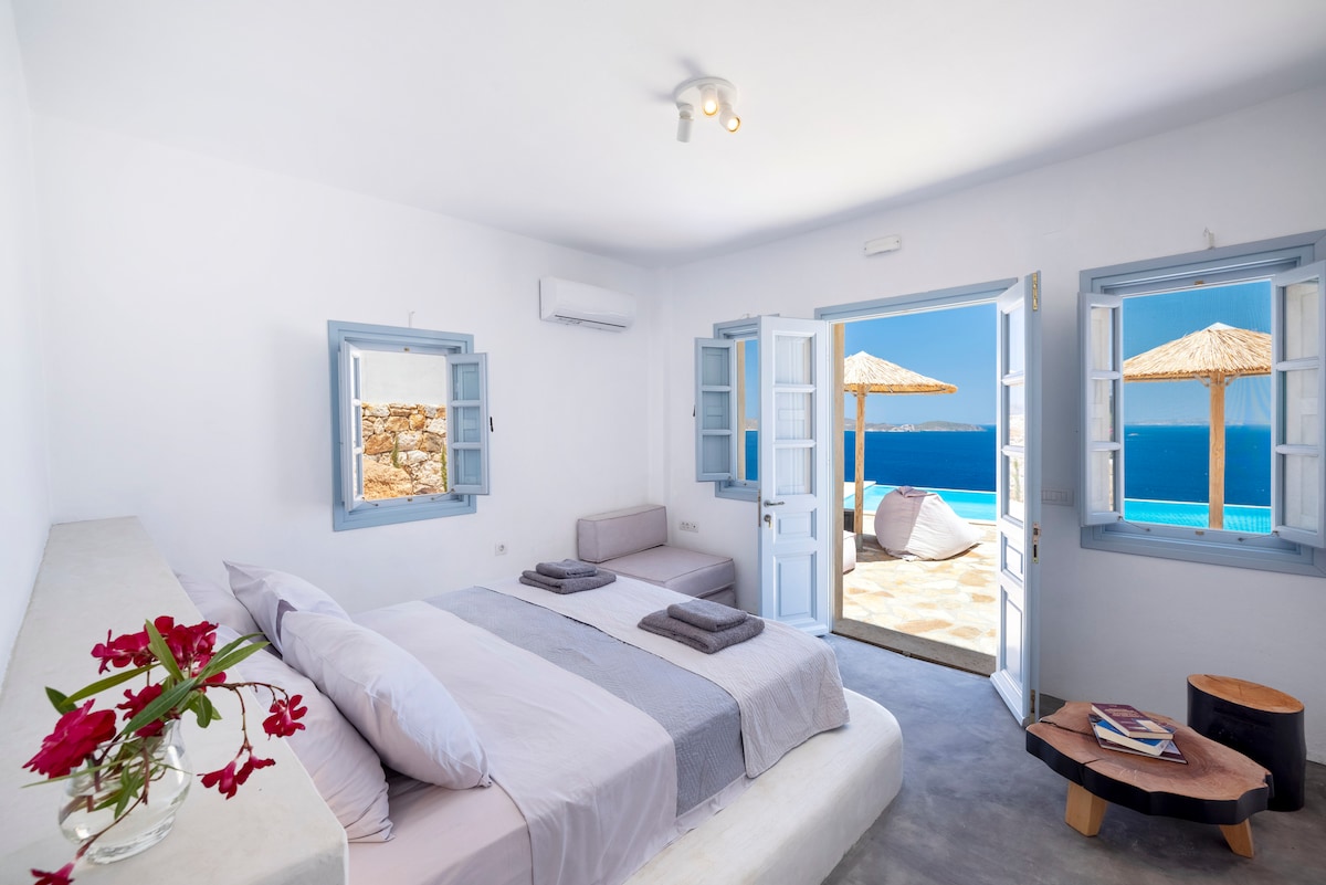 A serene bedroom is styled in light tones, featuring a spacious bed and a second smaller bed. Open shutters reveal breathtaking sea views and access to a sun terrace with umbrellas. Decorative flowers add a touch of color to the minimalistic decor.