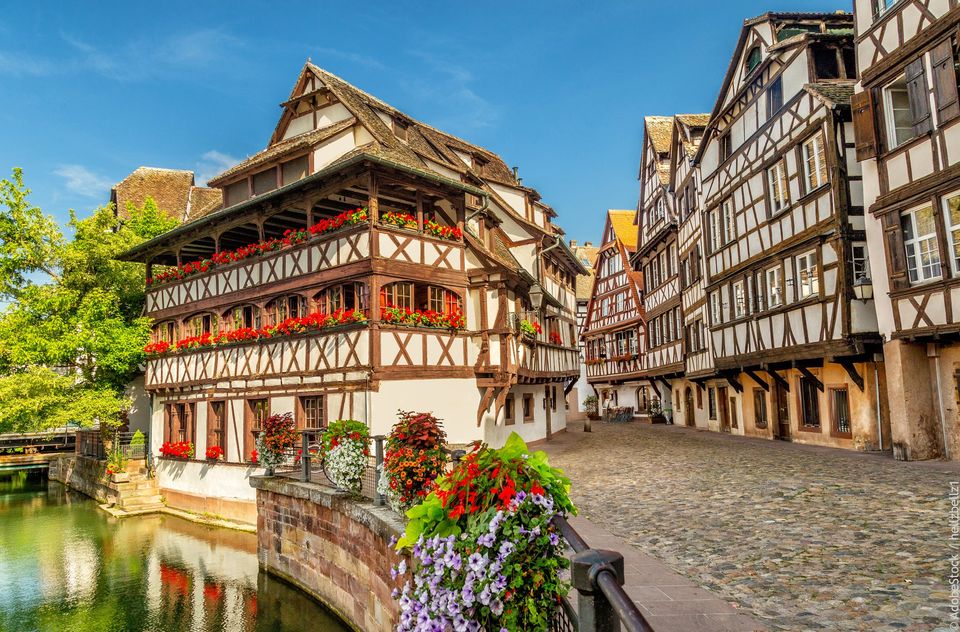 Historic timber-framed buildings line a picturesque cobblestone street. Colorful flower boxes adorn the windows, complementing the greenery along the canal. The tranquil water reflects the charming architecture, creating a serene environment in this lively Strasbourg neighborhood.