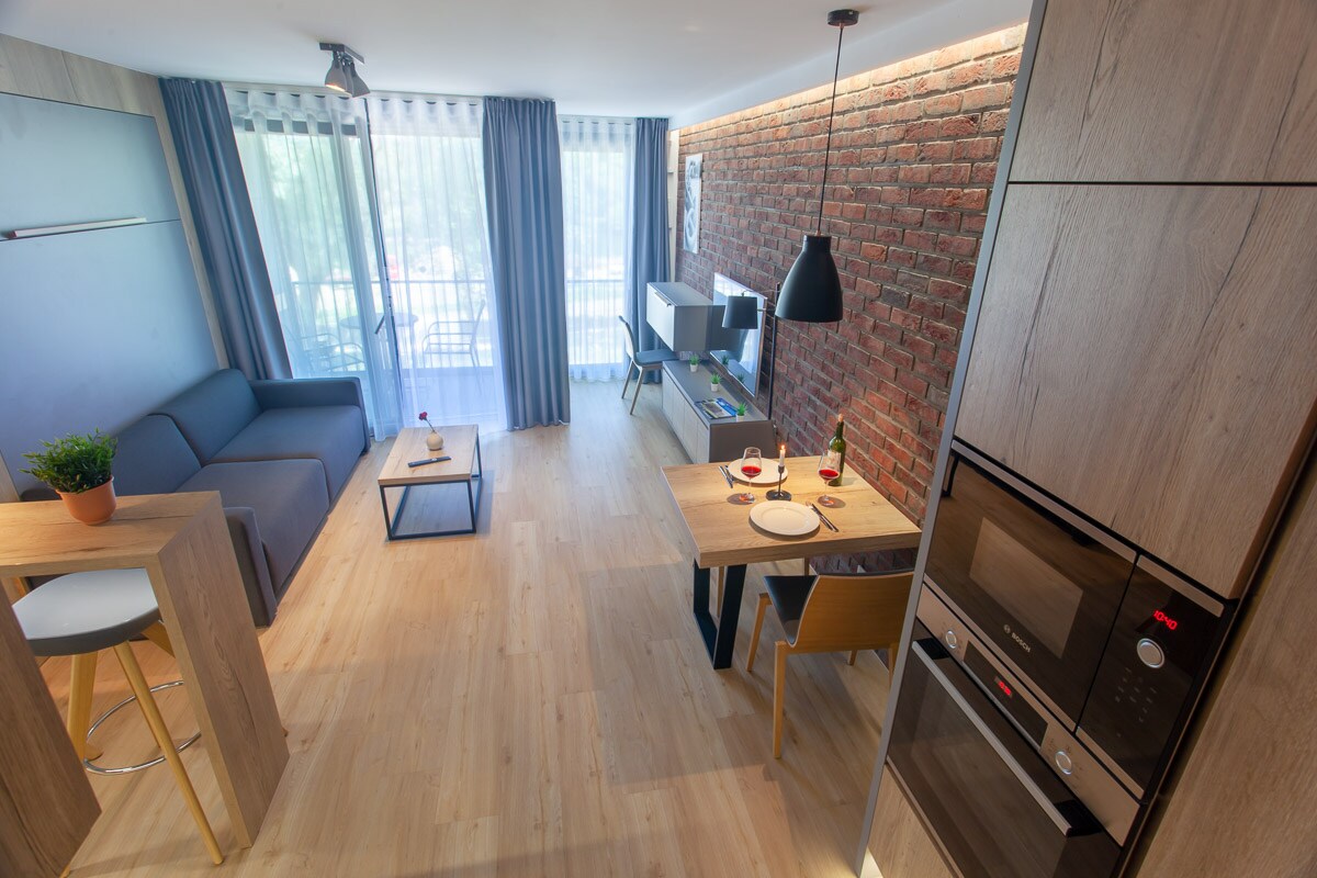 An open living area features a comfortable sofa and a dining table set for two. A small kitchenette is present, with modern appliances integrated into the cabinetry. Large glass doors provide natural light and lead to a balcony, with sheer curtains framing the view.