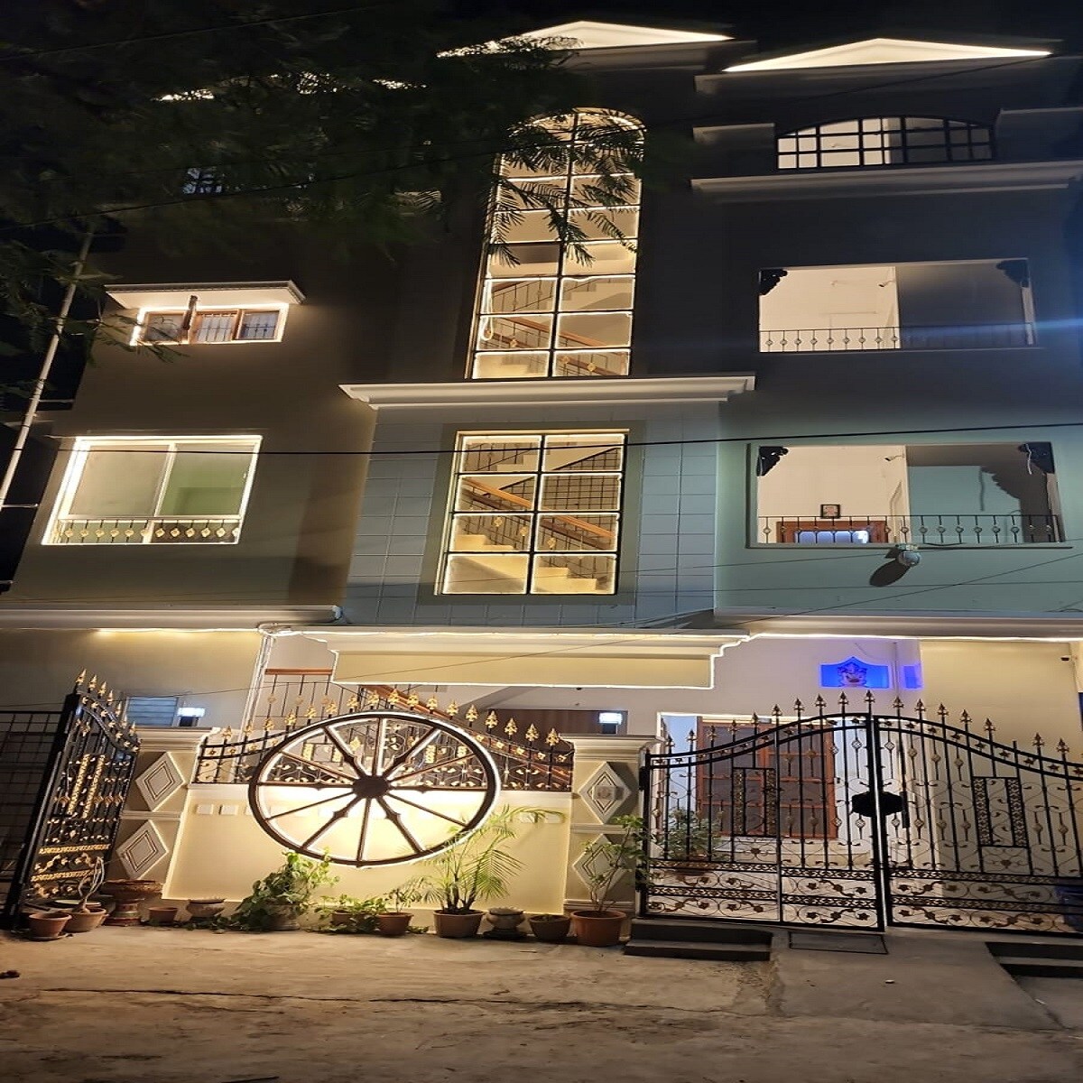 The exterior of a welcoming building is illuminated at night, showcasing modern architecture with large glass windows and ornate railings. A decorative wheel feature and potted plants enhance the entrance, which is secured by an elegant wrought iron gate.