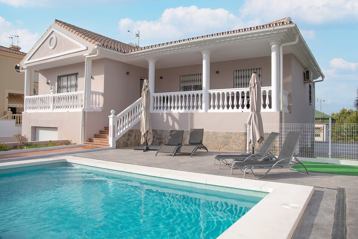 A modern villa features a spacious terrace with several lounge chairs placed beside a saltwater pool. The building showcases classic architectural details, including white columns and a tiled roof. Lush greenery surrounds the area, providing a peaceful outdoor setting.
