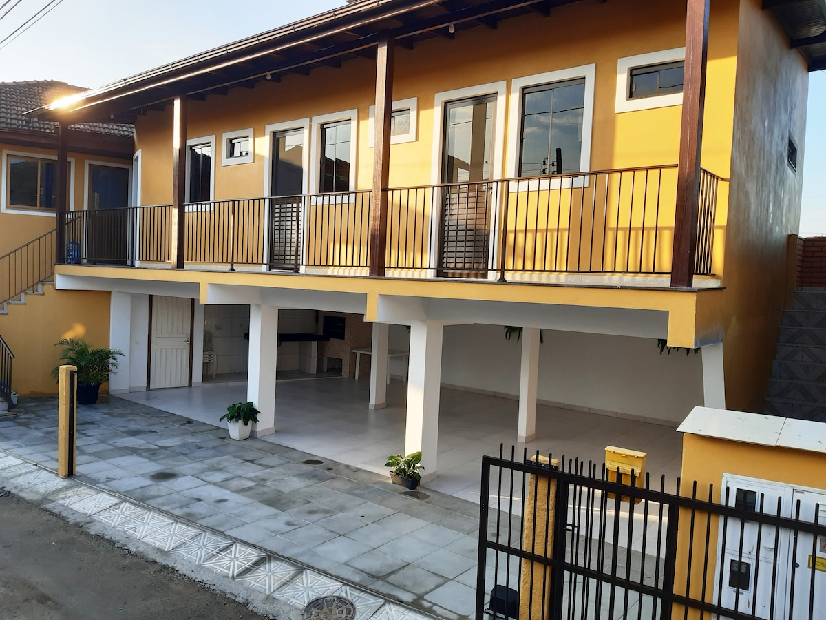 A two-story building features a brightly painted exterior in yellow, complemented by white accents. The upper level includes multiple large windows and a spacious balcony with railing. The ground floor offers an open space with a clean tiled surface, surrounded by a few potted plants.