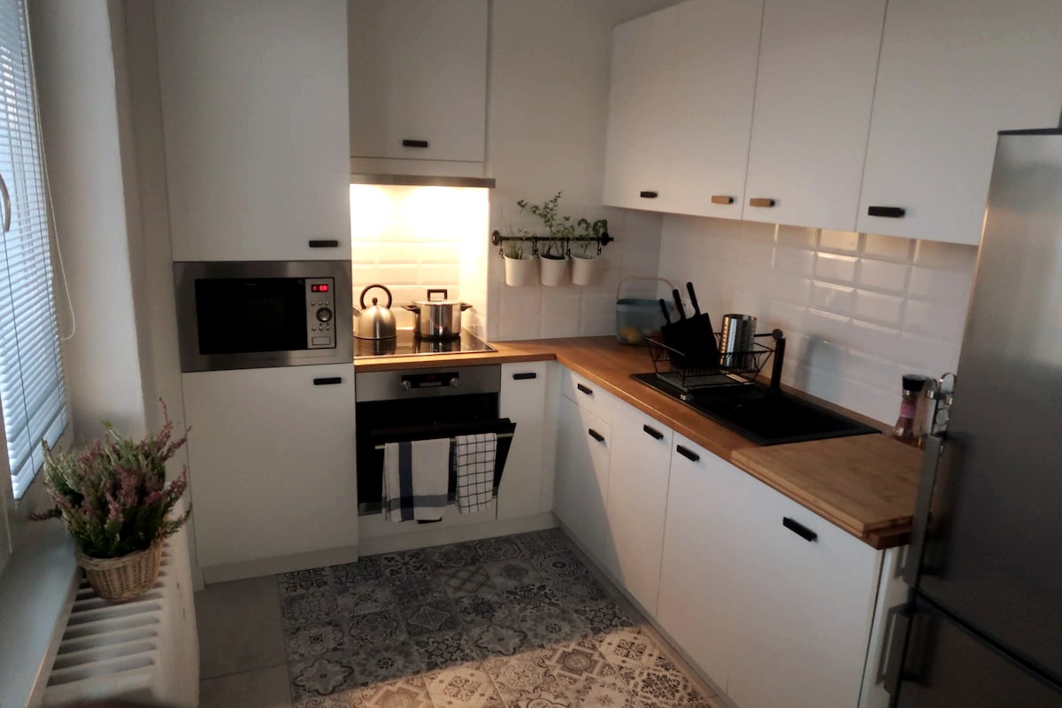 The kitchen is equipped with modern appliances, including a microwave and oven. Natural light filters through a window, illuminating the white cabinetry and wooden countertop. Various utensils are neatly organized, and potted plants add a touch of greenery to the space.