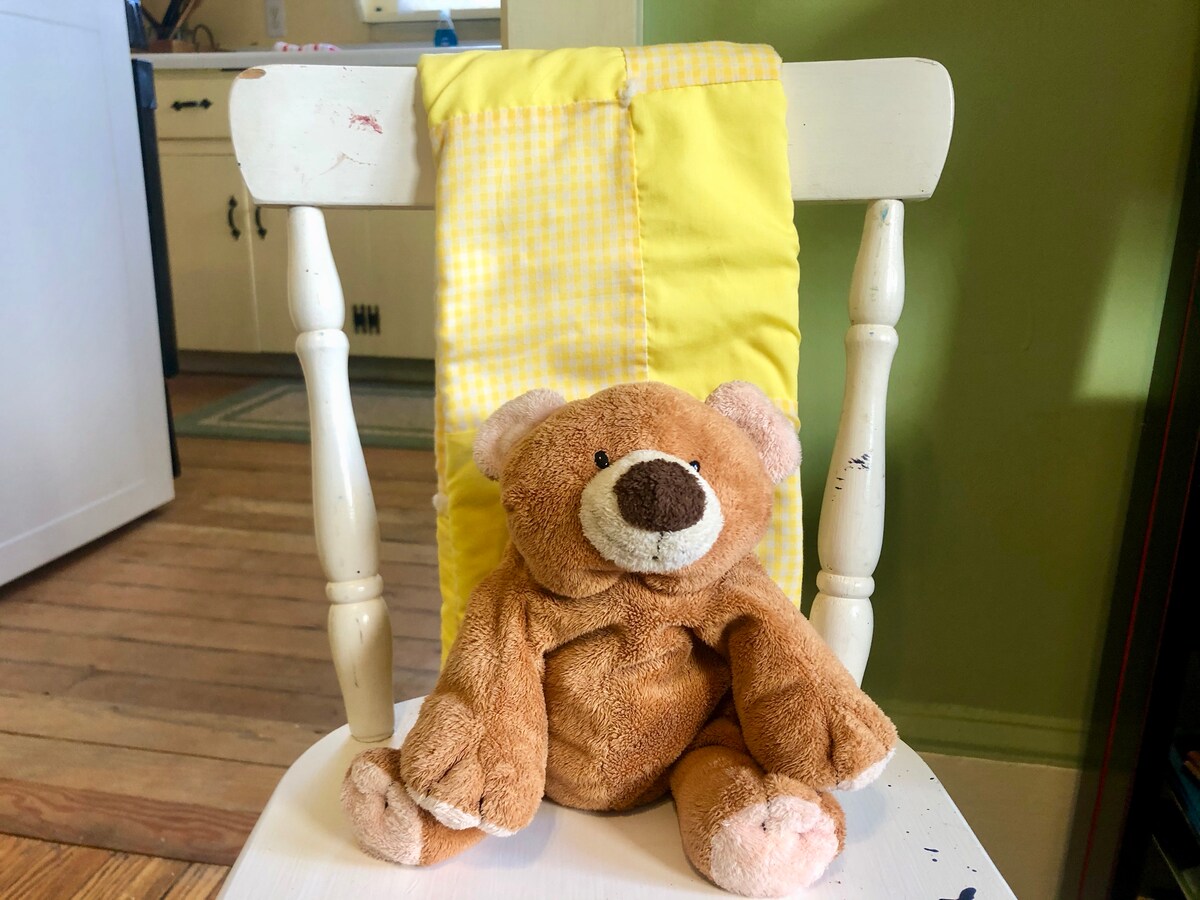 A soft, plush teddy bear is seated on a white wooden chair. The chair features a vibrant yellow fabric draped over the backrest. The hardwood floor and kitchen elements are subtly visible in the background.