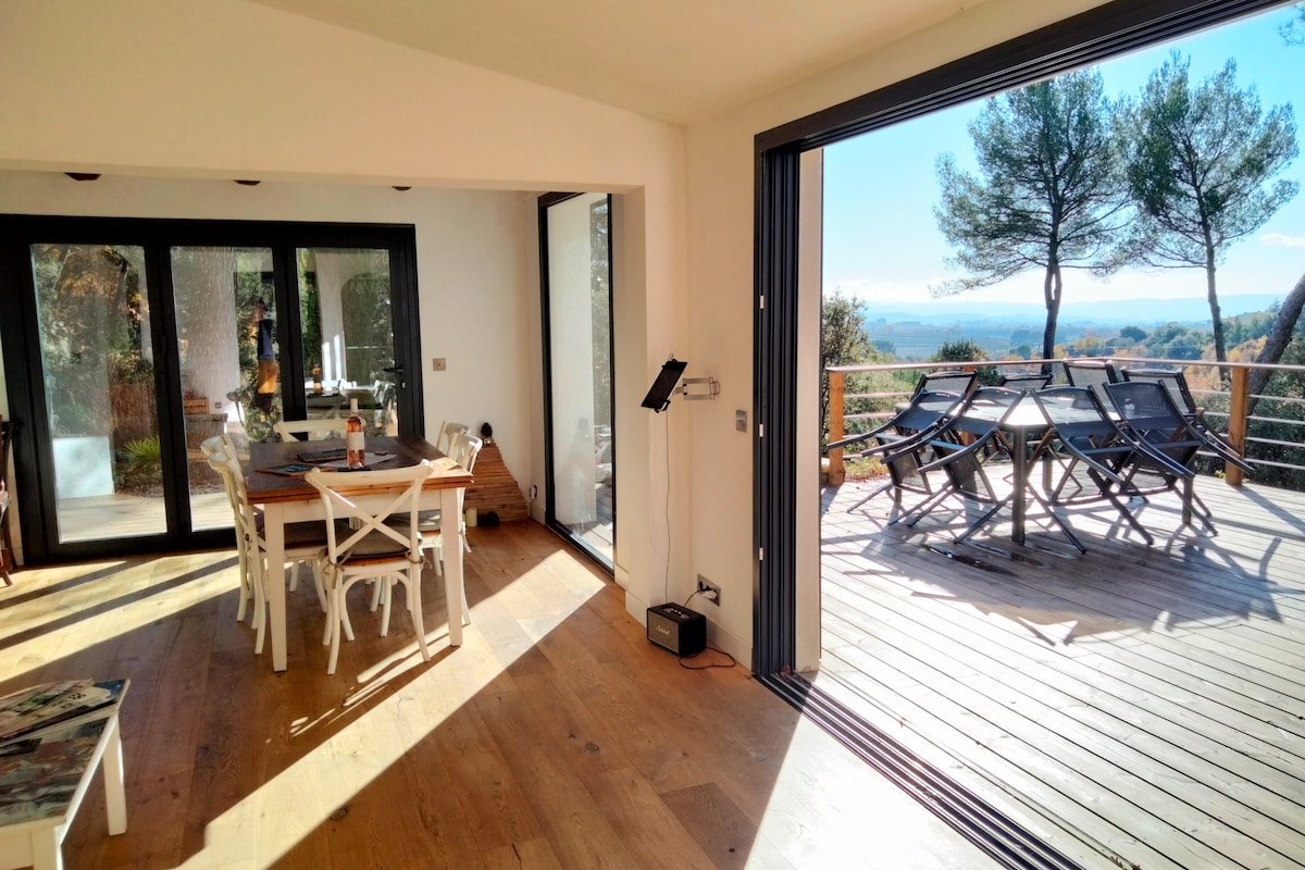 A bright dining area features a large table surrounded by white chairs, with large glass doors opening to a wooden terrace. The terrace showcases a dining set with chairs and a view of the surrounding nature and distant hills.
