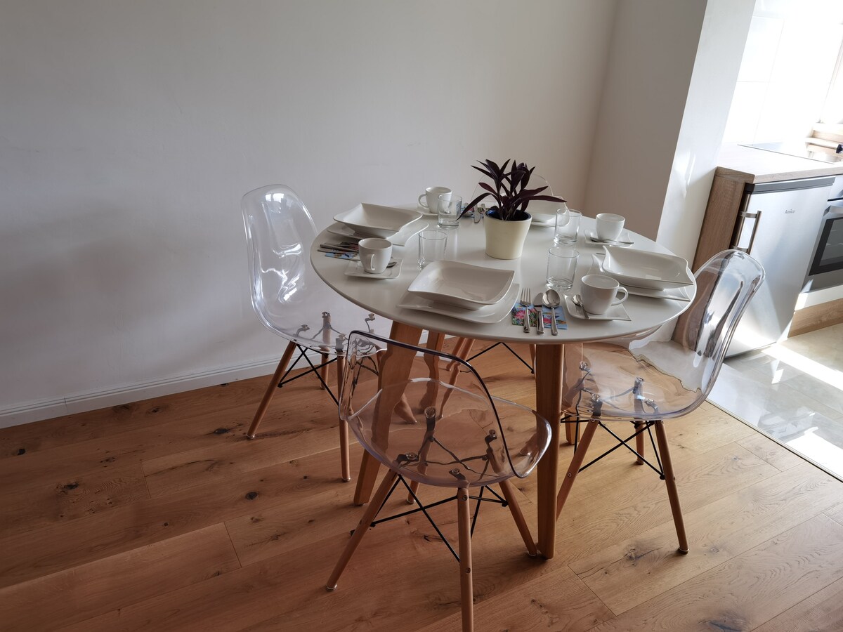 A round dining table is set for four, featuring white plates and cups, arranged neatly. Clear, modern chairs with wooden legs complement the clean design. A small potted plant sits in the center, with colorful markers positioned nearby. Natural light highlights the warm wooden floor.