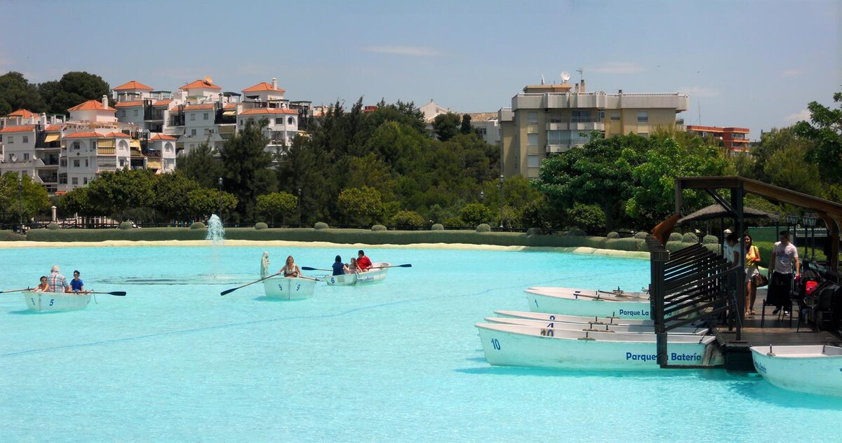 A serene lagoon is depicted with multiple rowboats gliding across the turquoise water, surrounded by lush green trees. In the background, residential buildings are visible, along with a small pavilion on the shore where visitors can be seen. A fountain adds a gentle touch to the peaceful scene.