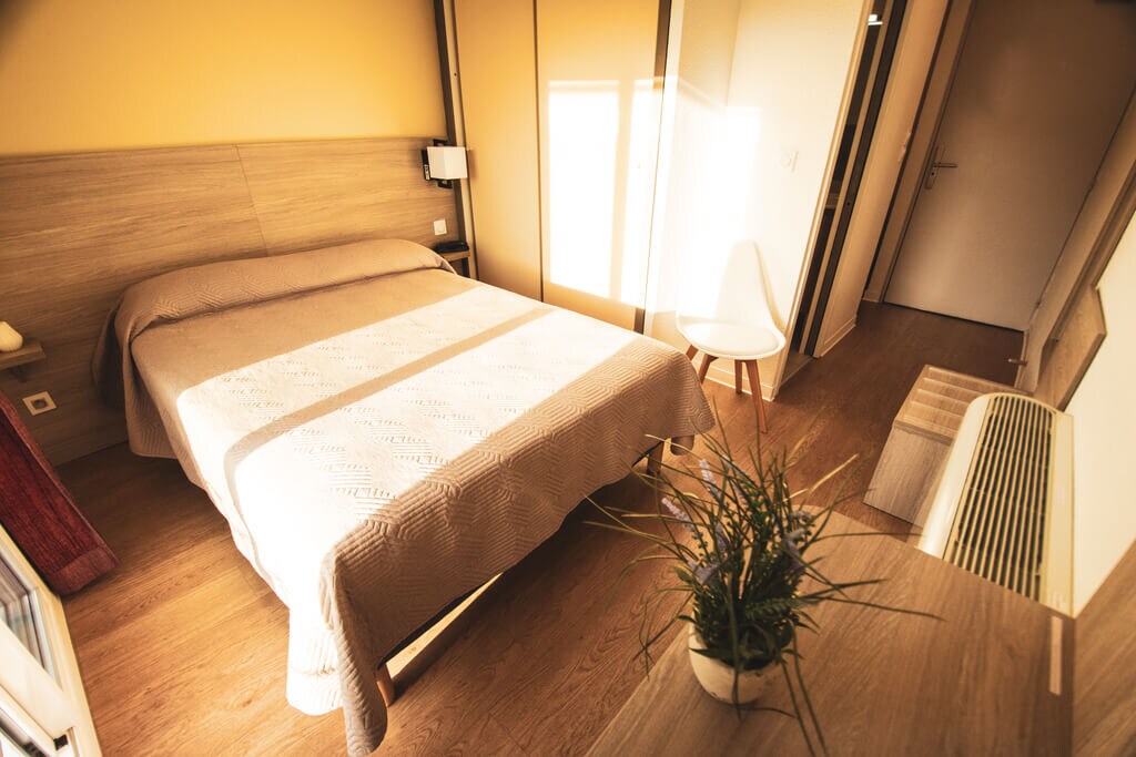 A room is featured with a spacious bed covered in a neutral-toned blanket. Natural light enters through large windows, illuminating the wooden flooring. A compact white chair and a small table are included in the decor, along with a potted plant adding a touch of greenery.