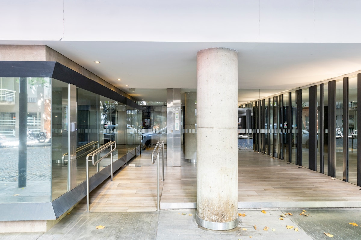 The entrance to the building features a spacious, modern design with glass walls and smooth wood flooring. A large concrete pillar is positioned centrally, while ramps for easy accessibility are visible on either side. Natural light floods the entryway, creating an inviting atmosphere.