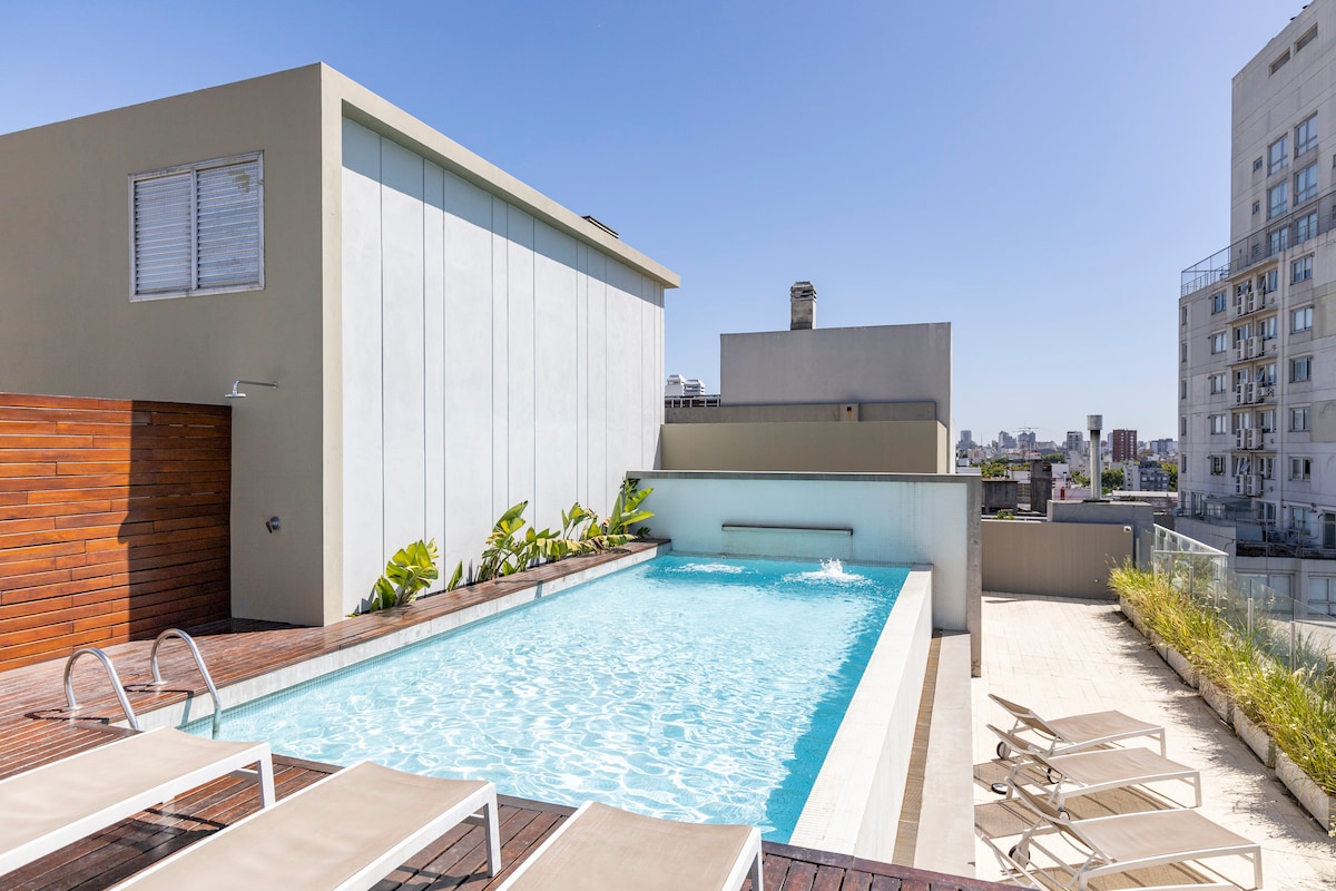 A rooftop pool area is showcased with a clear blue water surface reflecting the sky. Surrounding lounge chairs are arranged neatly on a wooden deck, complemented by tropical plants along the pool's edge. The building's modern facade provides a contemporary backdrop.