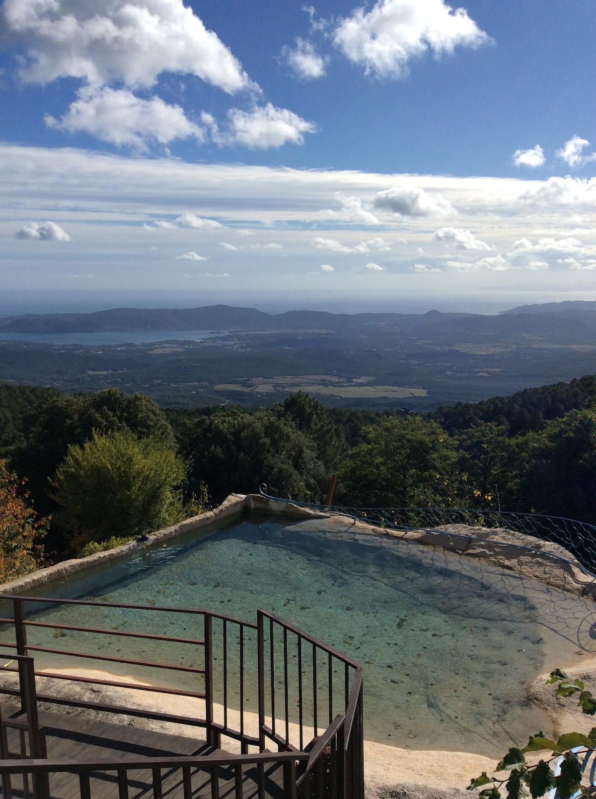 A serene outdoor pool is situated in a mountainous area, offering panoramic views of the landscape below. The water reflects soft sunlight, while surrounding trees provide a lush backdrop against a clear blue sky with scattered clouds.