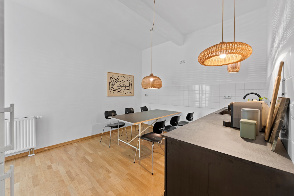 The dining area features a long table accompanied by six black chairs, providing ample space for gatherings. Natural light fills the room, highlighting the simple, modern décor. Unique pendant lights hang above the table, adding character to the inviting space.