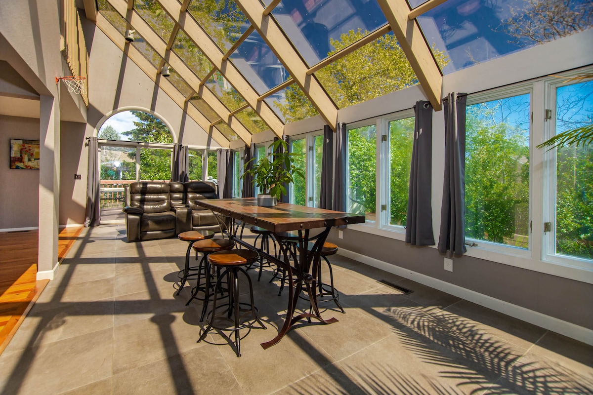 A bright solarium is filled with natural light from the glass roof and large windows overlooking greenery. A wooden dining table is positioned at the center, surrounded by stools. Comfortable seating is arranged in a cozy corner, inviting relaxation.