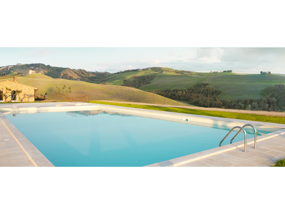 A seasonal outdoor pool is surrounded by lush green hills, offering a serene landscape. The pool's clear water reflects the sky, and the surrounding area features gentle slopes. A few steps lead into the water, providing easy access.