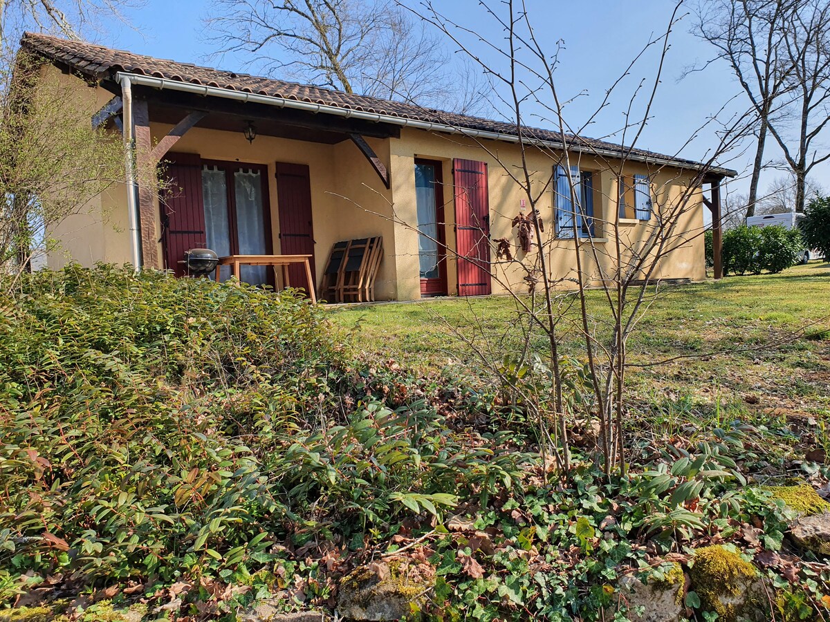 A single-story gîte is shown, featuring a warm exterior with reddish shutters. A covered terrace area is visible, furnished with chairs and a table. The surrounding garden displays lush greenery and low shrubs, enhancing the serene outdoor atmosphere.