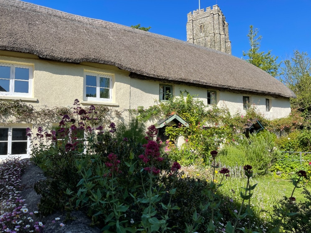 The charming cottage features a thatched roof and is set against a clear blue sky. Colorful flowers bloom in the well-kept garden, with the historic Norman church tower visible in the background, surrounded by greenery.