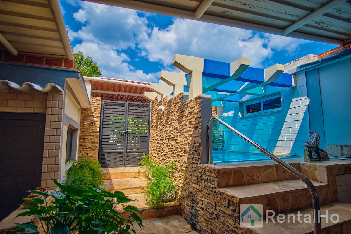 A serene outdoor area features a pool surrounded by natural stone walls and a well-maintained pathway leading to wooden gates. Shade is provided by blue awnings overhead, while greenery adds a touch of nature to the inviting space.