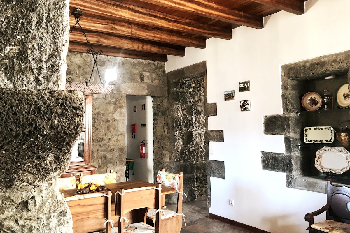 The interior showcases rustic stone walls paired with wooden beams overhead, providing an authentic ambiance. A wooden dining table is surrounded by chairs, while vintage wall decor adds character. Natural light enters through a nearby window, creating a warm and inviting space.