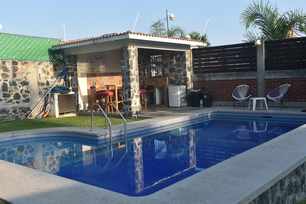 A private pool is surrounded by a well-maintained lawn. Next to the pool, a shaded palapa features outdoor seating and a built-in grill. The tranquil blue water reflects the clear sky, inviting relaxation and leisure activities.