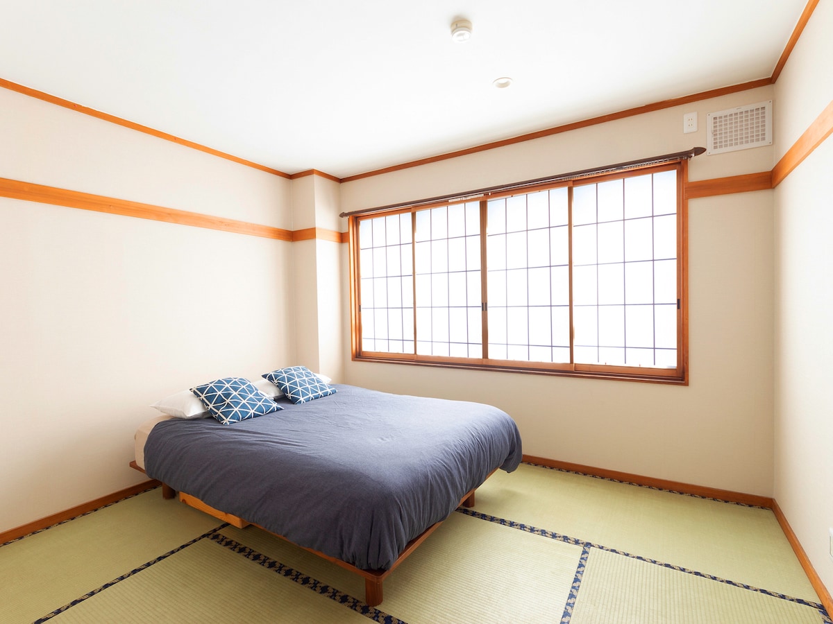 A spacious Japanese-style room features tatami flooring and a large futon positioned against a wall. Two cushions are displayed on the futon. A large window with shoji screens allows natural light to fill the room, enhancing the serene atmosphere.