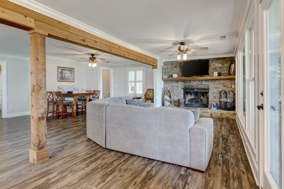 The spacious living area features a comfortable sectional sofa facing a stone fireplace. Two ceiling fans hang overhead, and a large television is mounted above the fireplace. A dining table with wooden chairs is visible in the background, providing an inviting gathering space.