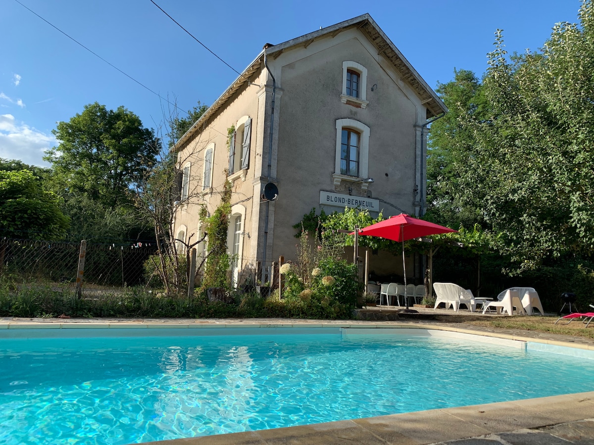 A tranquil private swimming pool is surrounded by a spacious outdoor area featuring lounge chairs and a shaded patio under a red umbrella. The charming station house can be seen in the background, framed by lush greenery and trees.