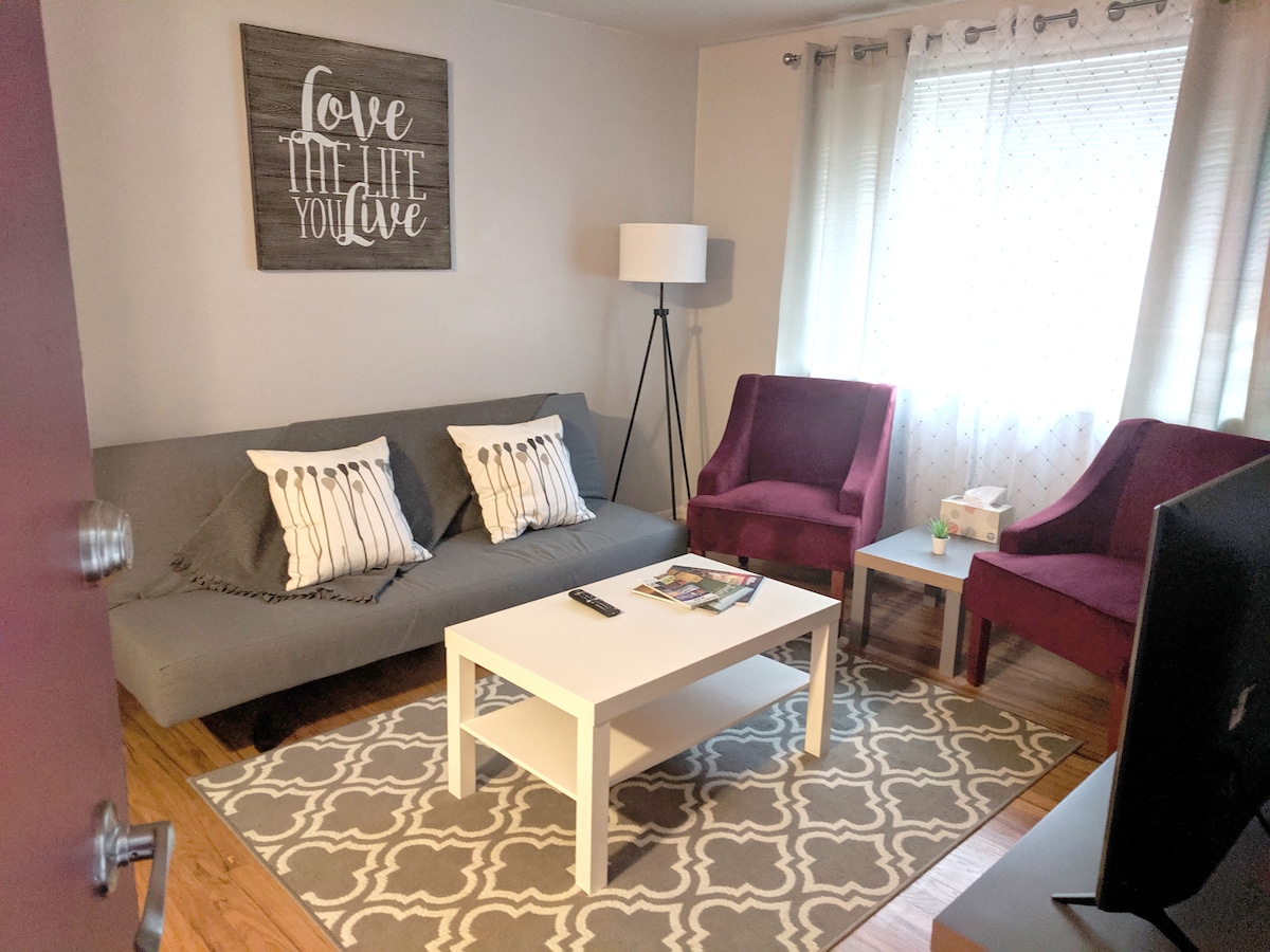 A welcoming living area displays a gray sofa with decorative pillows, complemented by two purple armchairs. A light rug with a geometric pattern covers the wooden floor, and a coffee table sits in the center. Natural light filters through sheer curtains, enhancing the space.