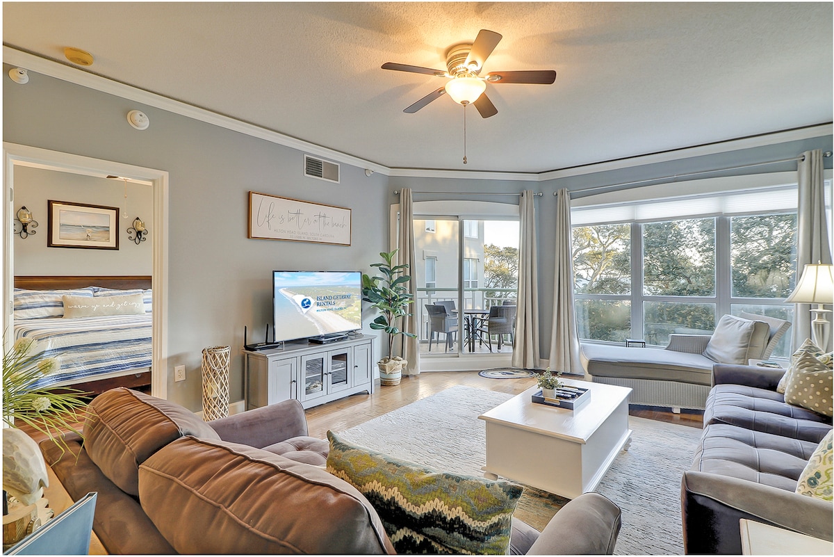 A bright and airy living area features comfortable seating options, including a gray sofa and armchairs. A large coffee table is centered on a soft area rug, with a television mounted on a light-colored media console. Floor-to-ceiling windows offer views of the surrounding landscape.