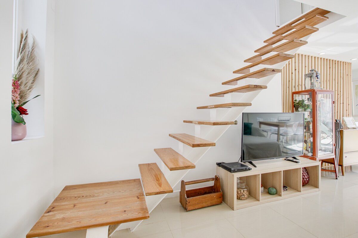 A modern staircase with wooden steps appears to float against a white wall, leading to an upper level. Below, a minimalist media console holds a television and decorative items. Natural light streams through a nearby window, illuminating the open space.