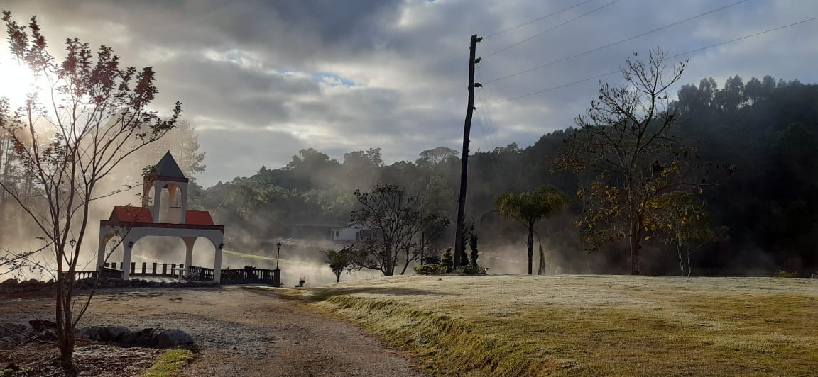 A scenic outdoor area features a gazebo with a red roof, surrounded by lush greenery and mist rising from the ground. Power lines run along the edge, while trees frame the distant view of a house, creating a serene and natural atmosphere.