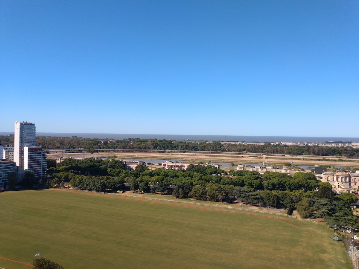 A panoramic view is presented, showcasing expansive greenery and open fields beneath a clear blue sky. In the distance, the cityscape and waterways can be seen, along with a glimpse of the racetrack, creating a vibrant and lively backdrop.