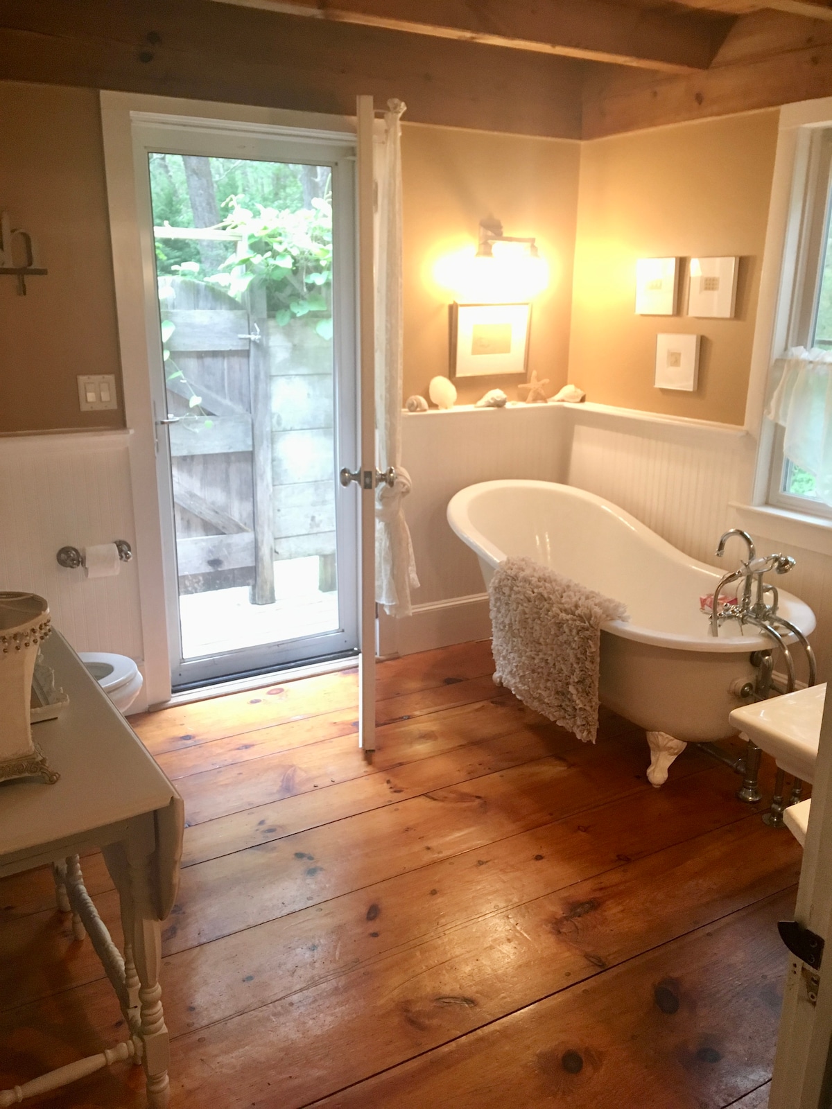 A bathroom features a vintage freestanding bathtub alongside a light-colored vanity. Natural wooden floors complement the rustic atmosphere, while soft lighting enhances the space. A glass door leads to an outdoor area framed by trees, providing a sense of connection to nature.