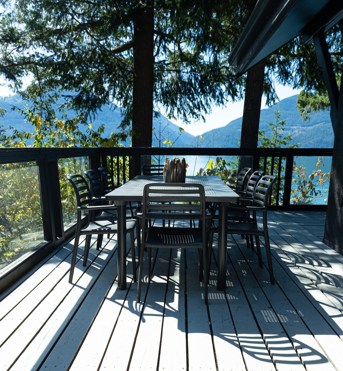 An outdoor dining area is featured on a wraparound porch, showcasing a large table surrounded by eight harmonious chairs. Tall trees provide shaded areas, and scenic mountain views are visible in the distance, creating a calming environment for dining.