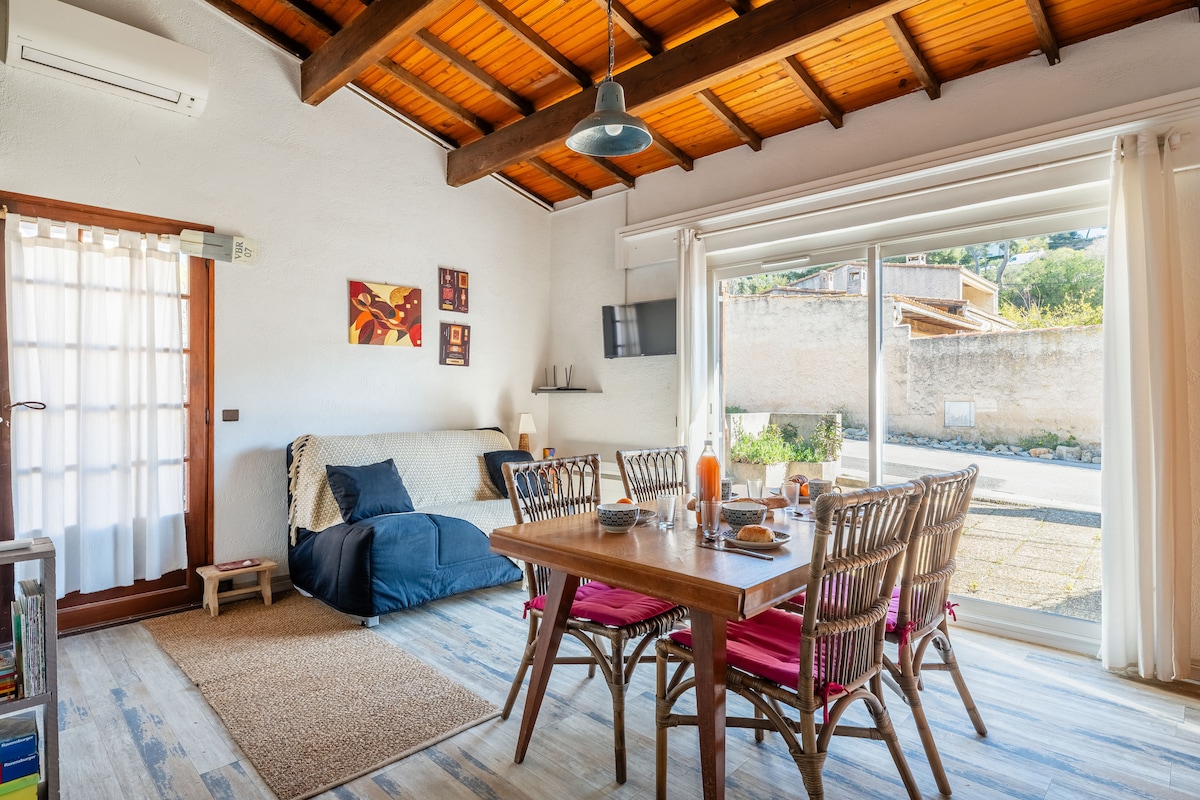 The living area showcases a spacious, bright environment with high ceilings and wooden beams. A convertible sofa is paired with a rustic dining table surrounded by woven chairs. Natural light floods in through large glass doors, which open to a serene outdoor view.