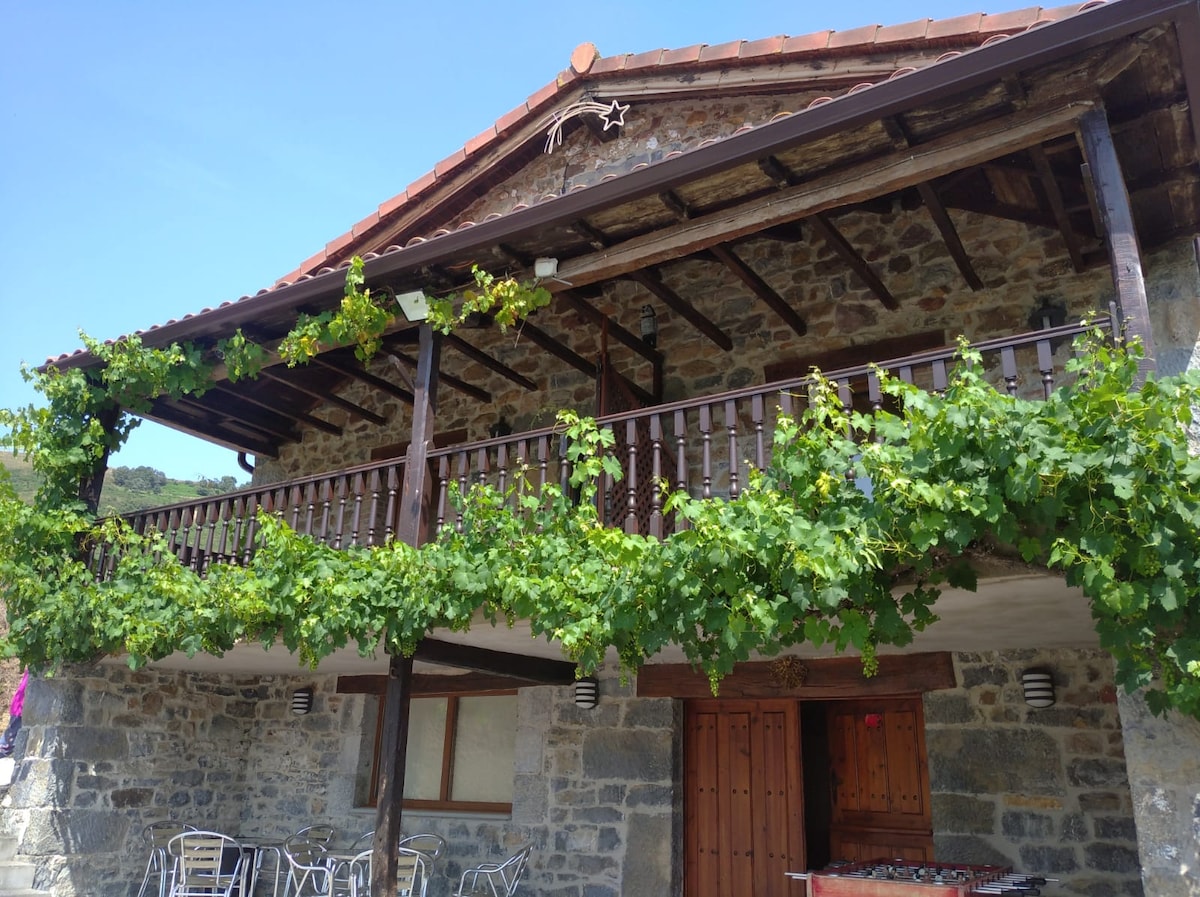 A rustic stone house with a wooden balcony, adorned with climbing vines. The architecture reflects the traditional mountain style, with a sloped roof and wooden beams. Large wooden doors provide access beneath the balcony, framed by natural stone walls.