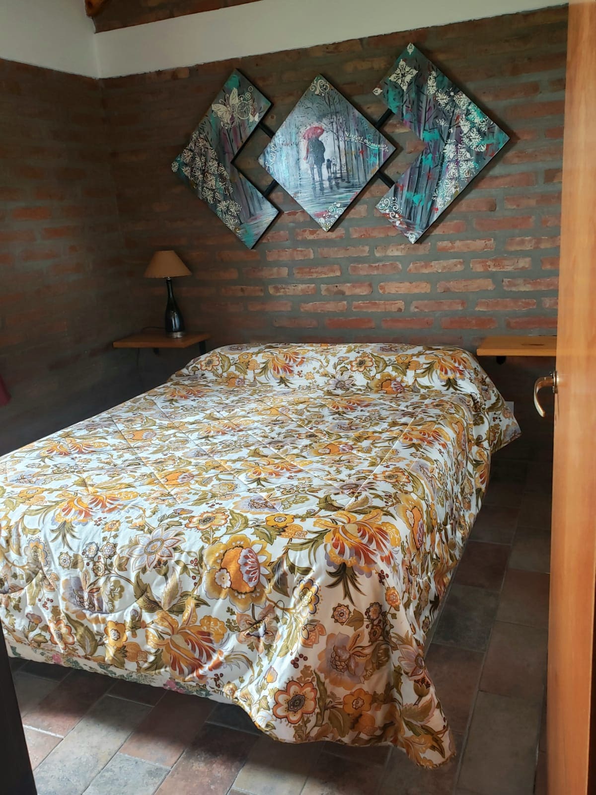 A sleeping area features a spacious bed covered with a floral patterned quilt. On the wall, three diamond-shaped artworks are displayed above the bed, and a bedside table with a lamp is positioned on one side. Exposed brick walls add a rustic touch.