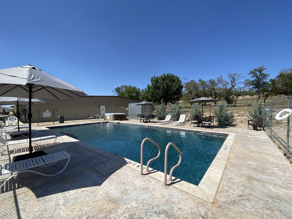 A fenced outdoor swimming pool is surrounded by lounge chairs and shaded areas under umbrellas. The clear blue water reflects the bright sky and is complemented by greenery in the background. A hot tub is positioned nearby, offering additional relaxation options.