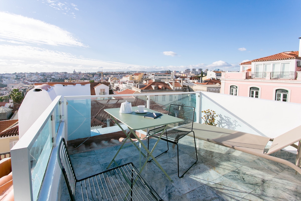 A spacious terrace offers panoramic views of the city and river, featuring a glass railing for safety. A metal dining table with matching chairs and a lounge chair are positioned on the outdoor space, providing a comfortable area for relaxation.