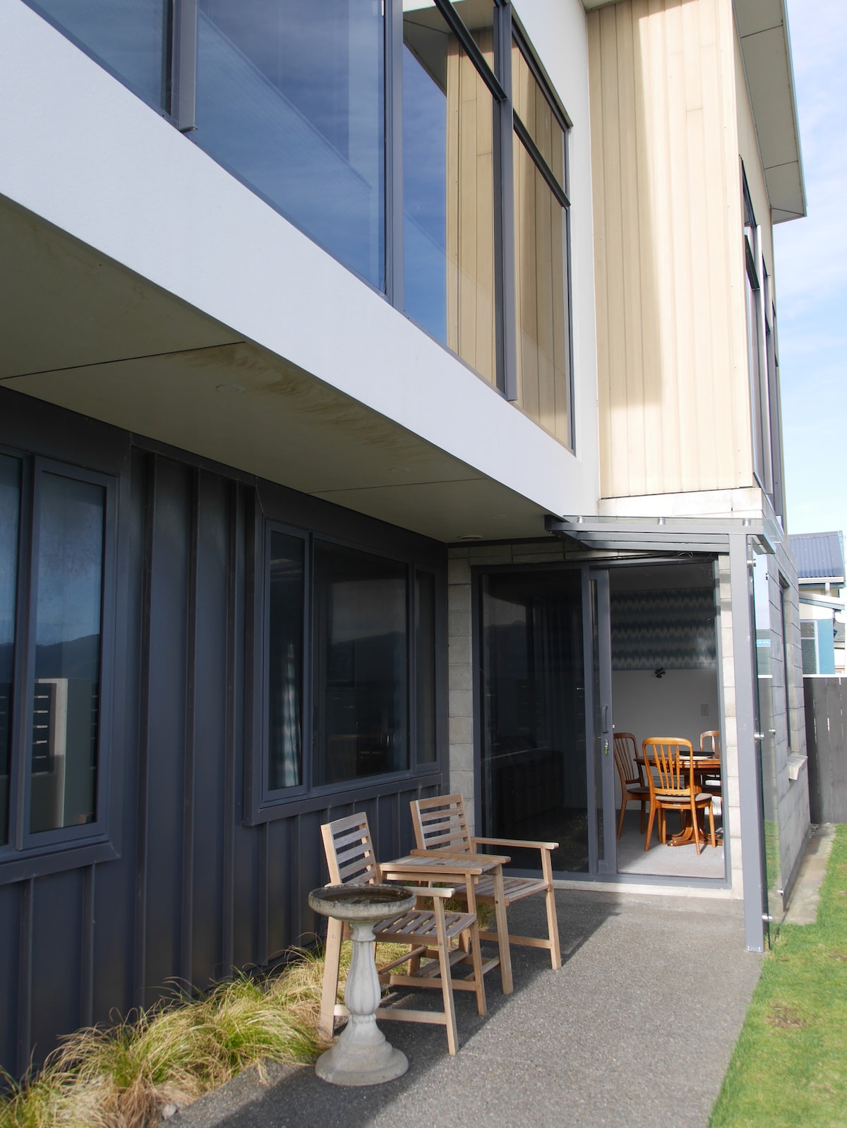 An outdoor area is depicted with two wooden chairs and a small table resting on a paved surface. Large windows provide a view into a dining area, allowing natural light to illuminate the interior. The exterior features a blend of materials, including wood and metal.