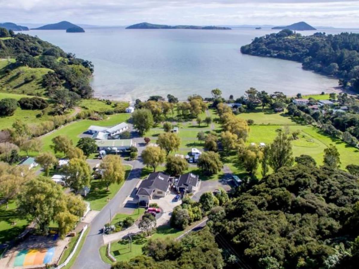 Aerial view captures the sprawling grounds of the Coromandel Shelly Beach TOP 10 Park, set against a backdrop of the bay. Lush greenery surrounds the area, with a variety of trees and pathways leading to the beach. Islands dot the horizon, contributing to the scenic coastal landscape.