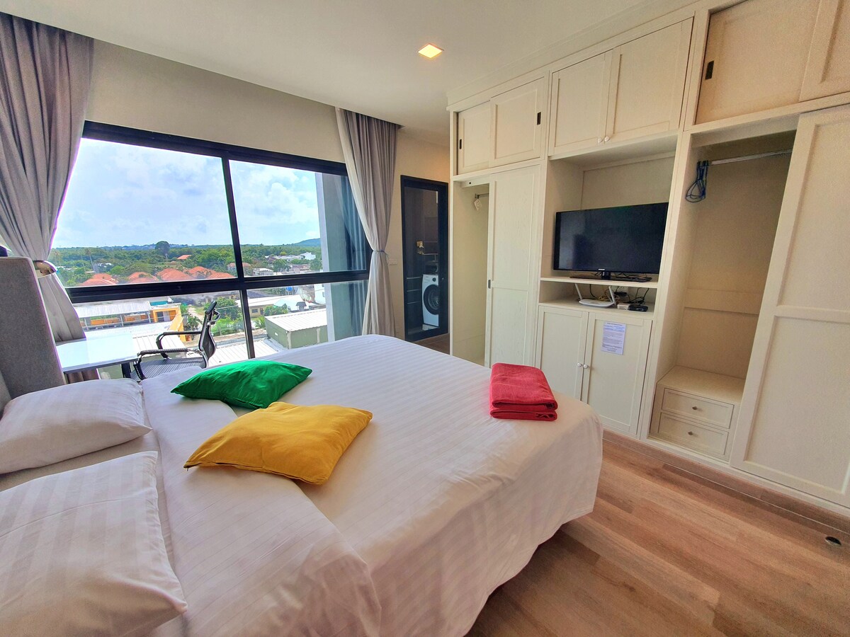 A studio room featuring a spacious bed adorned with colorful pillows. A television is mounted on the wall opposite the bed. Large windows offer a view of the outdoors, while soft curtains frame the space. Storage cabinets provide ample room for belongings.