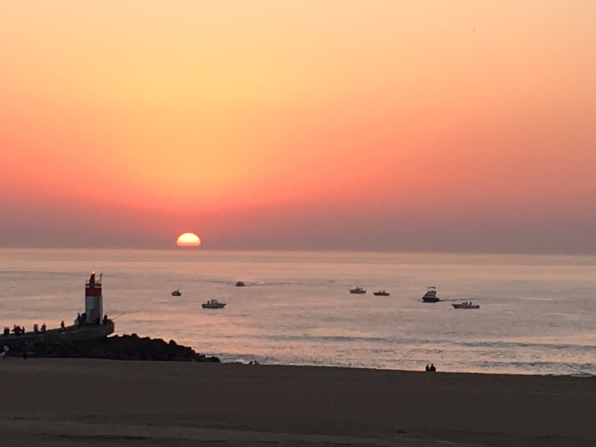 A serene sunset casts warm hues of orange and pink over the ocean. Silhouetted boats are seen gently floating on the water, while a lighthouse stands at the edge of the rocky shoreline. The horizon is tranquil, reflecting the fading sunlight.